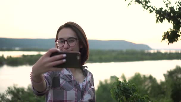 Egy fiatal nő áll egy dombon szemben a folyó és a hegyek hátterével, és fényképeket készít magáról. A lány szelfit készít a természetben..