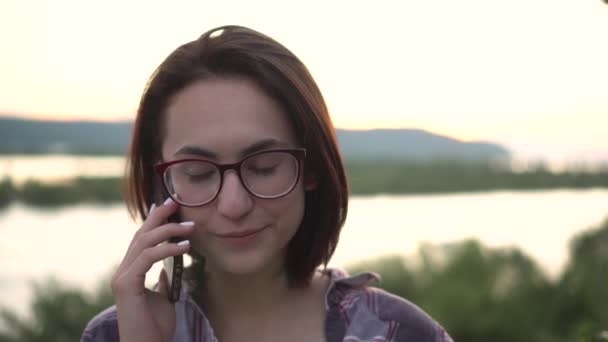 Молода жінка стоїть на пагорбі на тлі річки і гір і розмовляє по телефону. Дівчина розмовляє по телефону в природі крупним планом . — стокове відео
