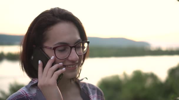 En ung kvinna står på en kulle mot bakgrunden av floden och bergen och talar i telefon. Flickan pratar i telefon i naturen närbild. — Stockvideo