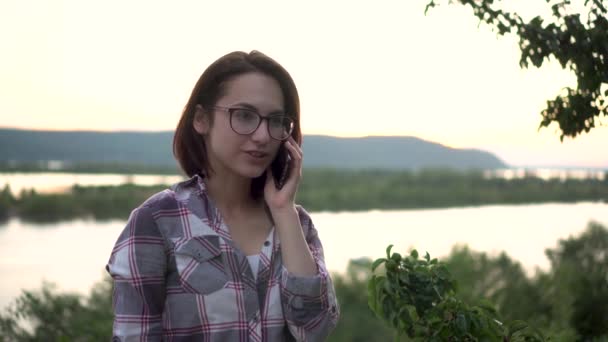 En ung kvinna står på en kulle mot bakgrunden av floden och bergen och talar i telefon. Flickan pratar i telefon i naturen närbild. — Stockvideo