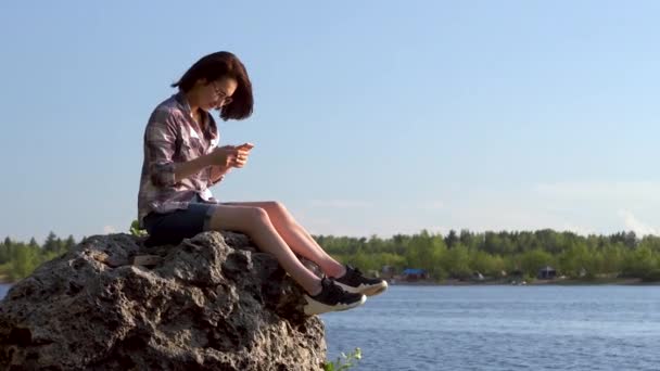 Μια νεαρή γυναίκα κάθεται σε μια πέτρα δίπλα στο ποτάμι με ένα τηλέφωνο στα χέρια της. Το κορίτσι στέλνει μηνύματα στο κοντινό κινητό.. — Αρχείο Βίντεο