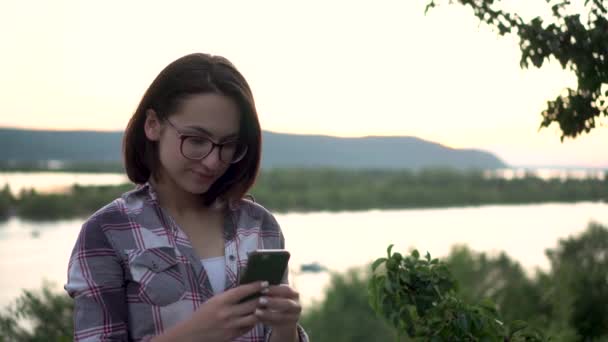 Une jeune femme se tient sur une colline sur le fond de la rivière et des montagnes avec un téléphone dans ses mains. La fille est dans la correspondance au téléphone dans la nature. — Video