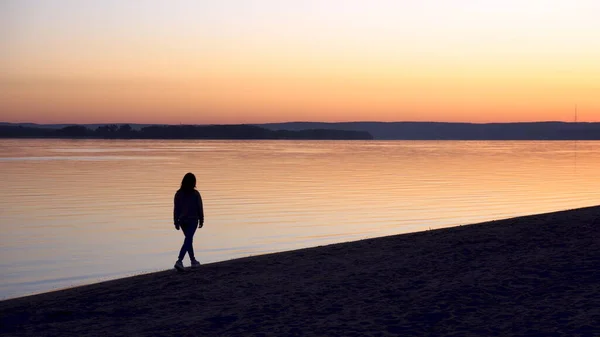 Mladá žena kráčí po pláži podél řeky za úsvitu. Silueta dívky. Jde vstříc úsvitu. — Stock fotografie