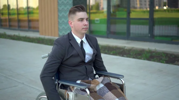 Jungunternehmer im Anzug im Rollstuhl. Ein ernsthafter Mann fährt im Rollstuhl gegen ein Geschäftszentrum. Spezielle Transportmittel für Behinderte. — Stockfoto