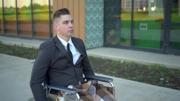 Joven hombre de negocios en traje en silla de ruedas. Un hombre serio monta en una silla de ruedas y mira su reloj contra el centro de negocios . —  Fotos de Stock
