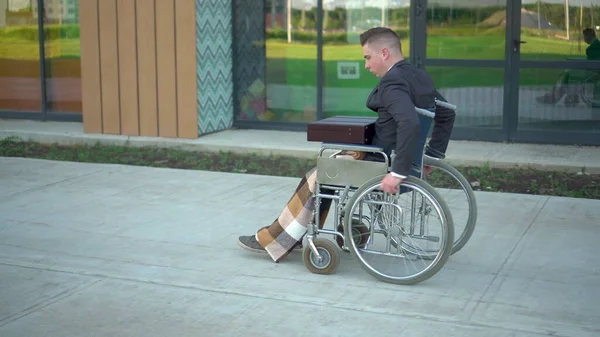 Jeune homme d'affaires en costume en fauteuil roulant. Un homme sérieux roule en fauteuil roulant contre un centre d'affaires. Transport spécial pour handicapés. — Photo