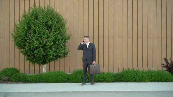 Молодой бизнесмен в костюме говорит по телефону. Серьезный человек, стоящий с портфелем в руке на заднем плане здания с клумбой. — стоковое фото