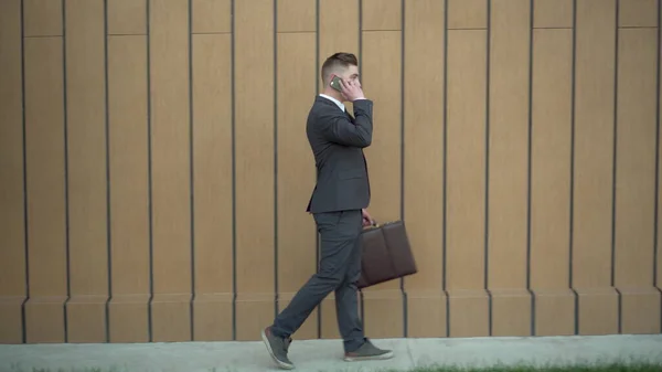 Молодий бізнесмен у костюмі говорить по телефону. Серйозна людина йде з портфелем в руці вздовж будівлі з телефоном . — стокове фото