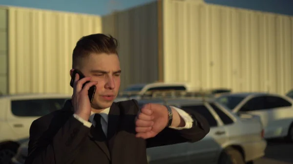 Um jovem empresário de fato fala ao telefone. Um homem sério está andando no estacionamento e olhando para seu relógio com um fecho de telefone . — Fotografia de Stock