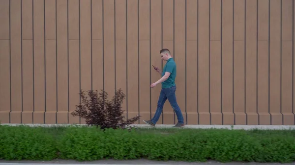 Молодой человек идет с телефоном в руке. Мужчина идет рядом со зданием и ведет переписку в смартфоне. — стоковое фото