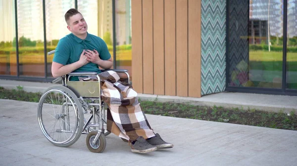 Un jeune homme conduit un fauteuil roulant et une crise cardiaque se produit. Un homme en fauteuil roulant sur le fond d'un bâtiment en verre saisit son cœur. Transport spécial pour handicapés. — Photo