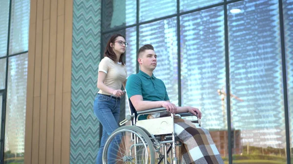 Uma jovem carrega um jovem numa cadeira de rodas. Um assistente carrega uma pessoa com deficiência em uma cadeira de rodas ao longo da rua. Transporte especial para deficientes . — Fotografia de Stock