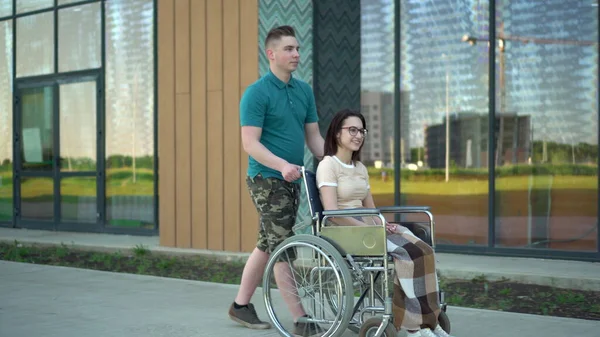 Egy fiatalember egy fiatal nőt cipel tolószékben. Egy asszisztens tolószékben cipel egy mozgássérültet az utcán. Speciális szállítás a fogyatékkal élők számára. — Stock Fotó