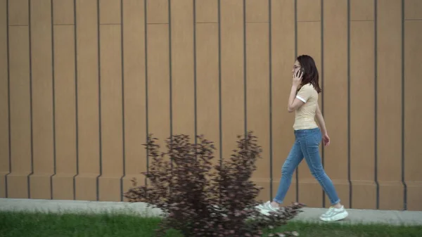 Una joven está caminando y hablando por teléfono. Una chica camina cerca del edificio y habla por teléfono. . —  Fotos de Stock