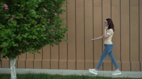 Una joven está caminando y hablando por teléfono. Una chica camina cerca del edificio y habla por teléfono. . —  Fotos de Stock