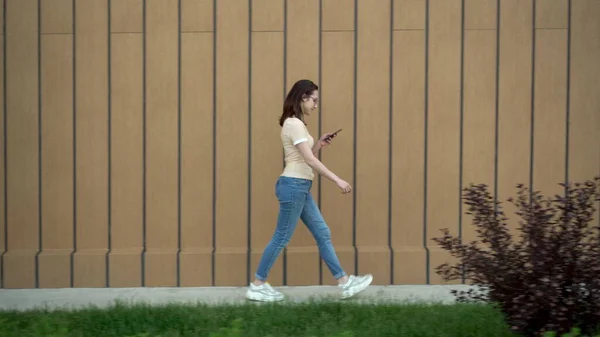 Una joven va con un teléfono en la mano. Una chica camina cerca del edificio y lleva la correspondencia en un teléfono inteligente . —  Fotos de Stock