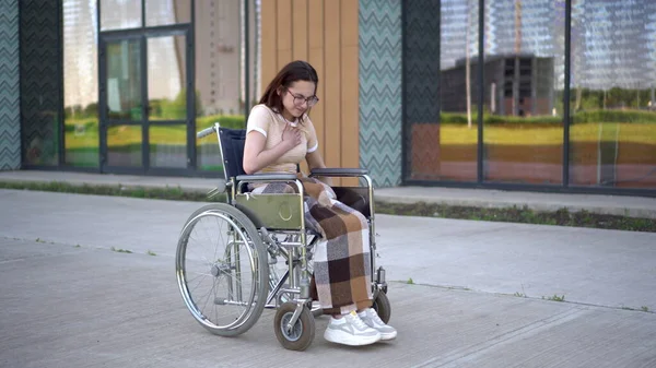 Uma jovem mulher monta uma cadeira de rodas e um ataque cardíaco ocorre. Uma menina em uma cadeira de rodas contra o fundo de um edifício de vidro agarra seu coração. Transporte especial para deficientes . — Fotografia de Stock