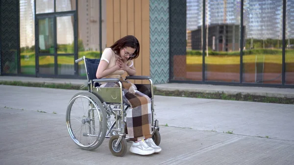 Uma jovem mulher monta uma cadeira de rodas e um ataque cardíaco ocorre. Uma menina em uma cadeira de rodas contra o fundo de um edifício de vidro agarra seu coração. Transporte especial para deficientes . — Fotografia de Stock