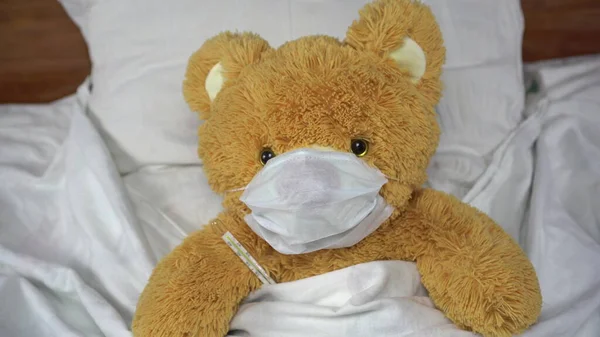 Miś mierzy temperaturę termometrem rtęciowym. Niedźwiedź leży w łóżku w masce medycznej.. — Zdjęcie stockowe