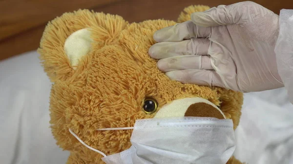 Teddy urso medir a temperatura com a mão closeup. O médico faz uma medição da temperatura aplicando uma mão em sua testa. O urso jaz na cama com uma máscara médica . — Fotografia de Stock