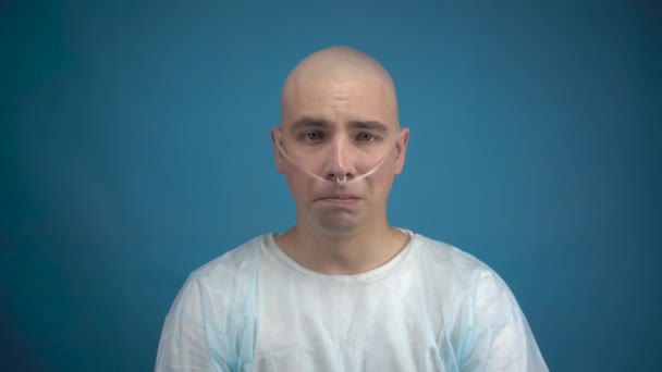 Łysy młody człowiek z onkologią patrzy w kamerę i płacze na niebieskim tle. Pacjent zakrywa twarz rękami. Utrata włosów w wyniku chemioterapii. — Wideo stockowe