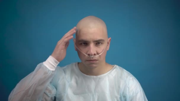 Łysy młody człowiek z onkologią niestety patrzy w obiektyw na niebieskim tle. Pacjent dotyka łysej głowy. Utrata włosów w wyniku chemioterapii. — Wideo stockowe