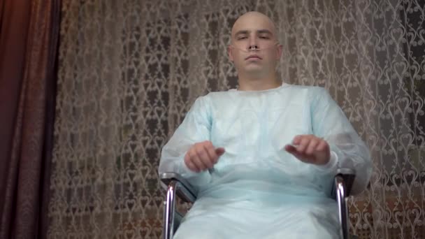 Un giovane calvo con il cancro guarda la telecamera e prega. Il paziente ha incrociato le braccia e prega con un sorriso a casa su una sedia a rotelle. Perdita di capelli a causa della chemioterapia. — Video Stock
