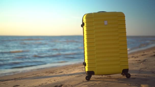 Żółta walizka stoi na plaży na tle morza. Podróżna walizka. Młoda kobieta w białej sukience biegnie w tle. — Wideo stockowe