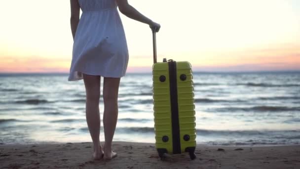 Молода жінка з жовтою валізою на пляжі біля моря. Дівчина в білій сукні дивиться на захід сонця крупним планом. Подорожі в теплі країни . — стокове відео