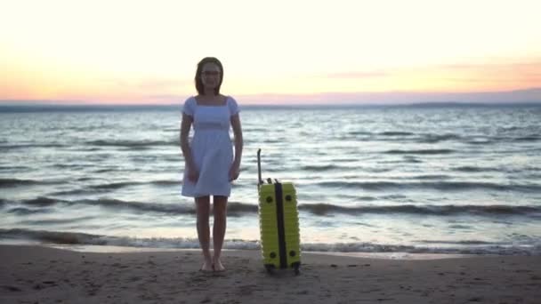 Mujer joven con una maleta amarilla en la playa junto al mar. Una niña en un vestido blanco junto al mar al atardecer levanta las manos con alegría. Viaje a países cálidos. — Vídeos de Stock