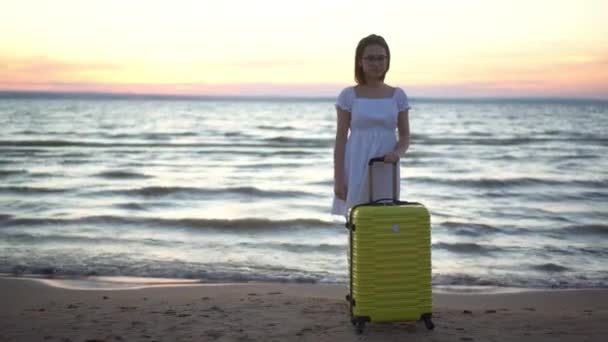 Молода жінка з жовтою валізою на пляжі біля моря. Дівчина в білій сукні на заході сонця. Подорожі в теплі країни . — стокове відео