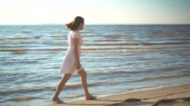 Çekici genç bir kadın deniz sahilinde yürüyor. Beyaz elbiseli bir kız sahilde yalınayak yürüyor..