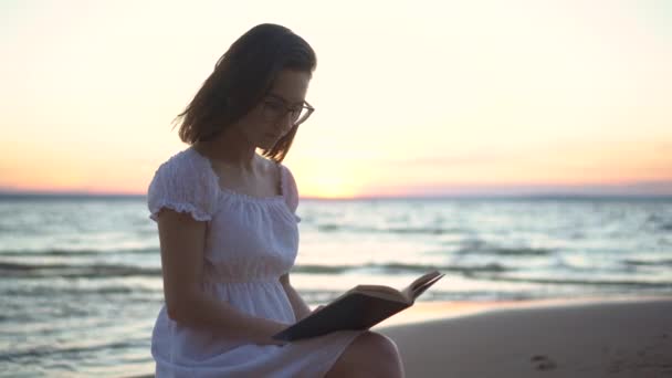 En ung kvinna sitter på en sten på stranden vid havet med en bok i händerna. En flicka i vit klänning vid solnedgången läser en bok närbild. — Stockvideo