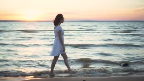 Çekici genç bir kadın gün batımında deniz sahilinde yürüyor. Beyaz elbiseli bir kız sahilde yalınayak yürüyor.. — Stok video