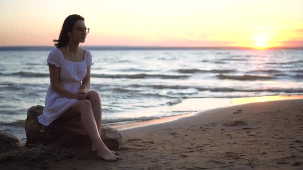 Een jonge vrouw zit op een steen op het strand aan zee met een koptelefoon in haar oren. Een meisje in een witte jurk bij zonsondergang luistert naar muziek op de hoofdtelefoon. — Stockvideo