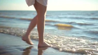 Genç bir kadın kumlu deniz kıyısında yalınayak duruyor. Kız ayaklarını ıslatıyor..