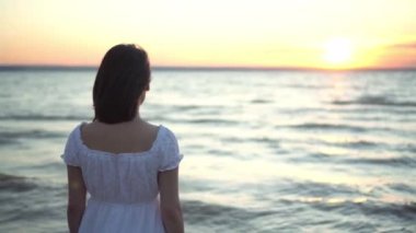 Çekici genç kadın deniz kenarında gün batımını seyrediyor. Beyaz elbiseli kız arkasında duruyor..