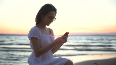 Genç bir kadın sahilde bir taşın üzerinde oturuyor. Elinde telefon var. Gün batımında beyaz elbiseli bir kız akıllı telefondan konuşuyor..