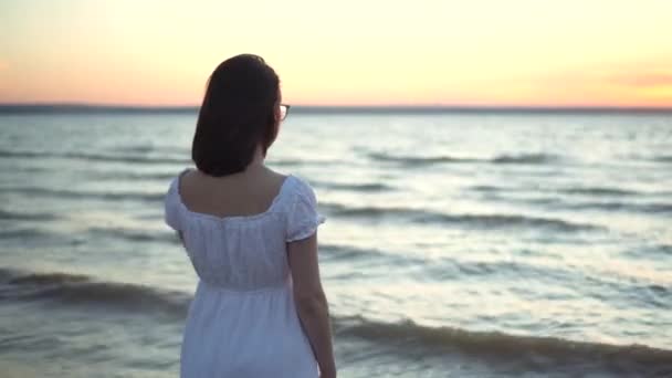 Atraktivní mladá žena se dívá na západ slunce na pláži u moře. Dívka v bílých šatech stojí zády.. — Stock video
