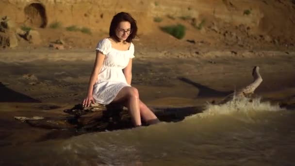 Eine attraktive junge Frau sitzt auf einem Baumstamm am Fluss. Ein Mädchen im weißen Kleid plätschert mit den Beinen im Fluss vor dem Hintergrund eines sandigen Hügels. — Stockvideo