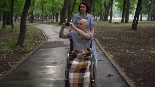 Žena vede mladého muže s onkologií na vozíčku přes park. Muž sedí s telefonem v ruce a fotí okolní přírodu. Ztráta vlasů v důsledku chemoterapie. — Stock video