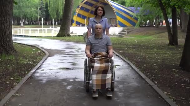 Une femme conduit un jeune homme avec oncologie dans un fauteuil roulant à travers le parc. Cet homme est chauve à cause de la chimiothérapie.. — Video