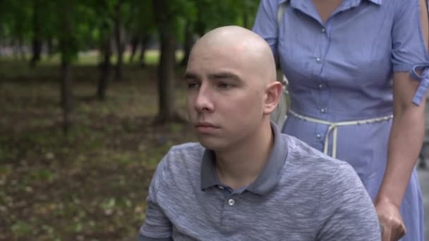 Жінка веде молодого чоловіка з онкологією в інвалідному візку через парк крупним планом. Чоловік лисий через хіміотерапію . — стокове відео