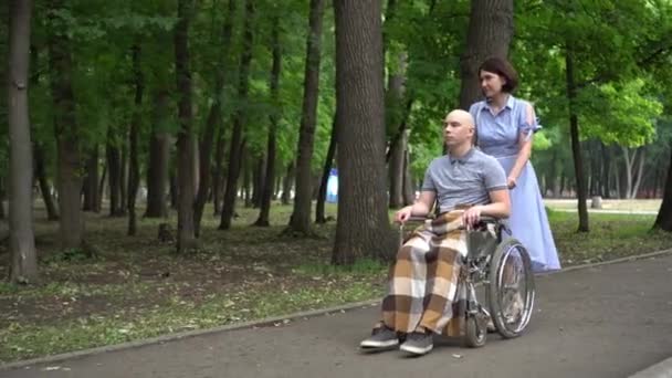 Žena vozí mladého muže s onkologií na vozíčku přes park. Ten muž je plešatý kvůli chemoterapii.. — Stock video