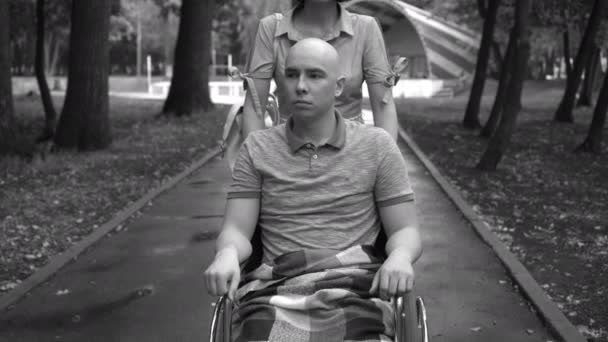 En kvinna kör en ung man med onkologi i rullstol genom parken. Mannen är skallig på grund av kemoterapi. Monokrom färg. — Stockvideo