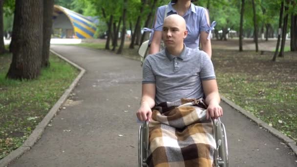 Eine Frau fährt einen jungen Mann mit Onkologie im Rollstuhl durch den Park. Der Mann hat wegen Chemotherapie eine Glatze. — Stockvideo