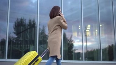 Genç bir kadın sarı bir bavulla yürüyor ve telefonda konuşuyor. Gözlüklü ve ceketli bir kız aynalı bina boyunca yürüyor..