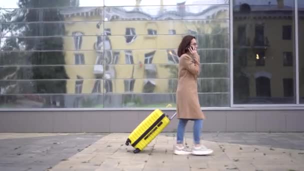 Mladá žena jde se žlutým kufříkem a mluví po telefonu. Dívka s brýlemi a kabátem kráčí po zrcadlové budově. — Stock video