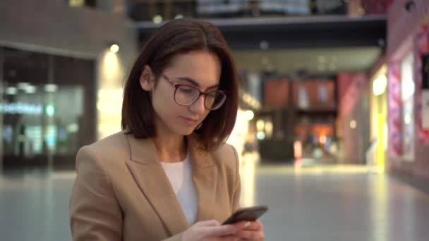 Una mujer joven se sienta con un teléfono en sus manos en un centro comercial con una gran palidez. Una chica con un abrigo rosa está enviando mensajes por teléfono. — Vídeo de stock