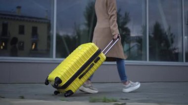 Genç bir kadın sarı bir bavulla yürüyor. Gözlüklü ve ceketli bir kız aynalı bina boyunca yürüyor..